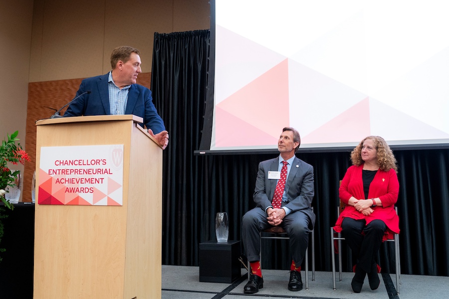 Mark Bakken speaks at a podium during the awards ceremony with Jennifer Mnookin and Charles Hoslet sit off to the side